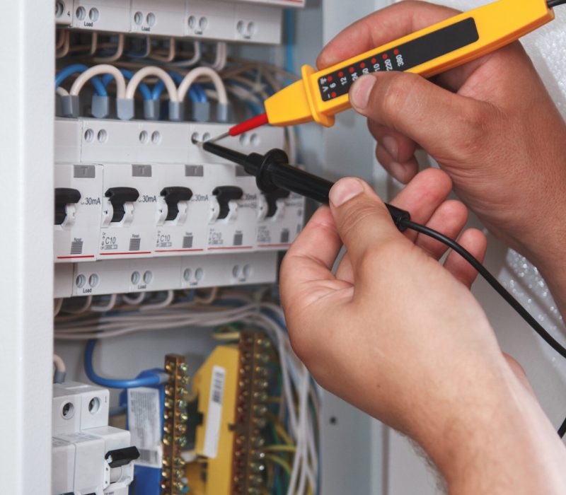 Hands of electrician with multimeter probe at an electrical switchgear cabinet examining fuse box
